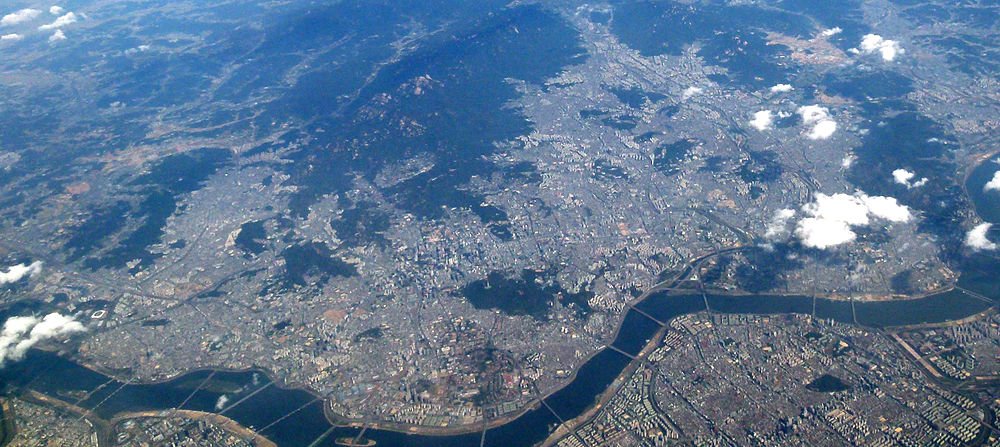 Aerial view of a large urban city with rivers, bridges, and scattered clouds over mountainous terrain