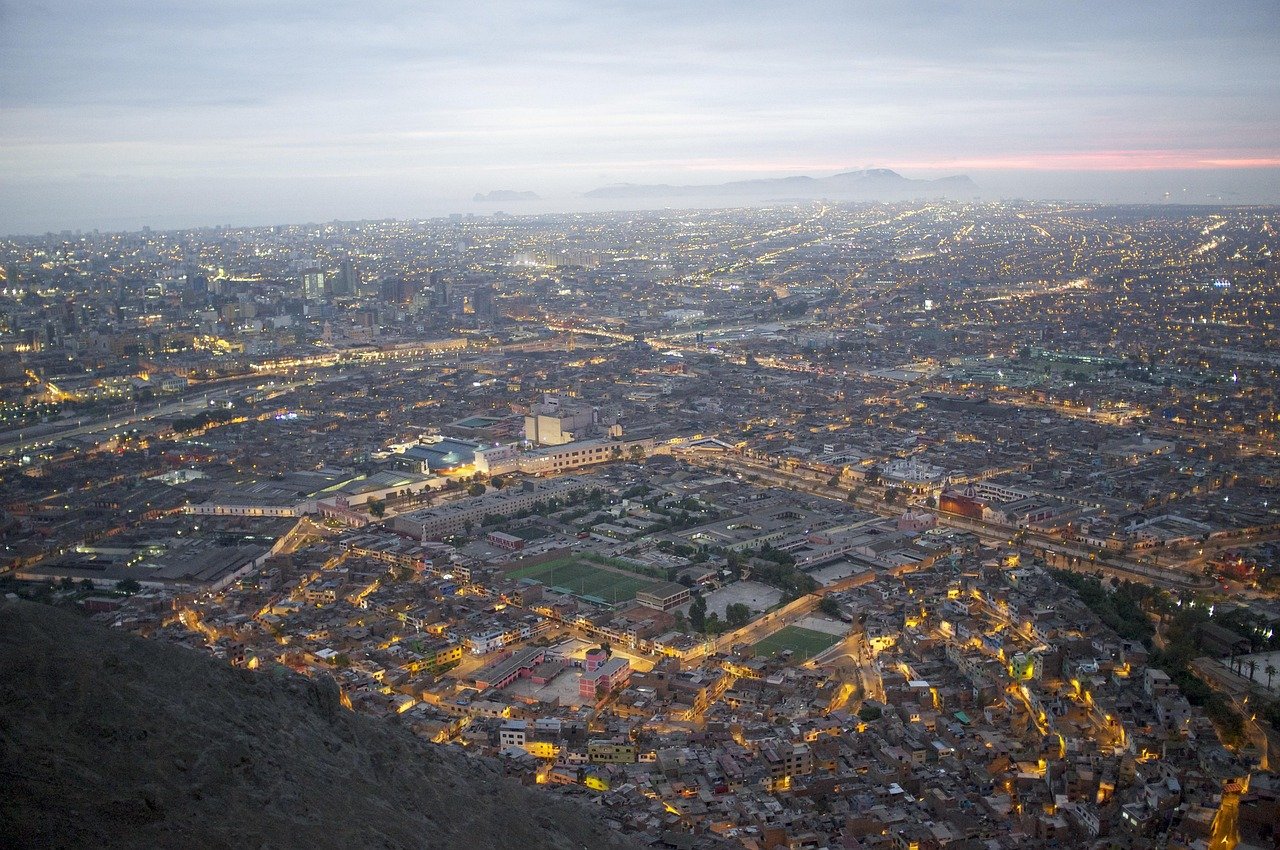 Aerial view of a sprawling cityscape at dusk with illuminated streets and distant mountains under a cloudy sky