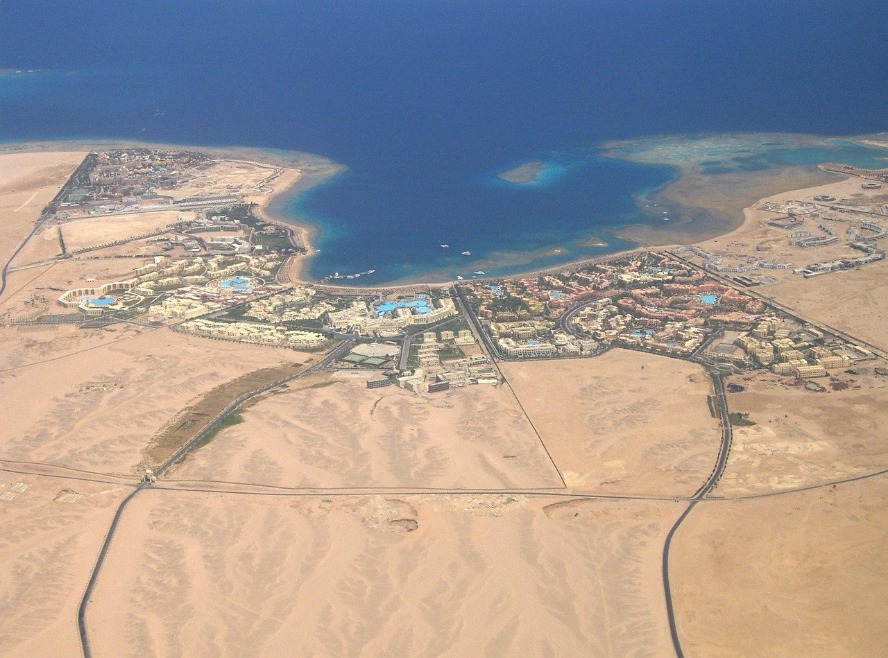 Aerial view of a coastal desert resort town with sandy terrain, blue sea, and clustered buildings with swimming pools