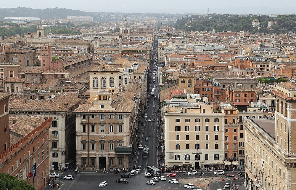 Aerial view of a busy street lined with historic buildings and cars in a European city with distant hills and greenery