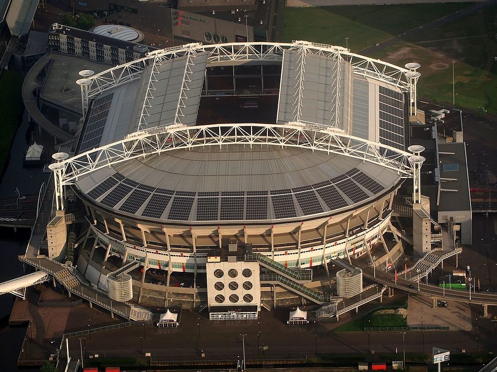 Aerial view of a large modern stadium with a partially open roof and solar panels on the roof structure