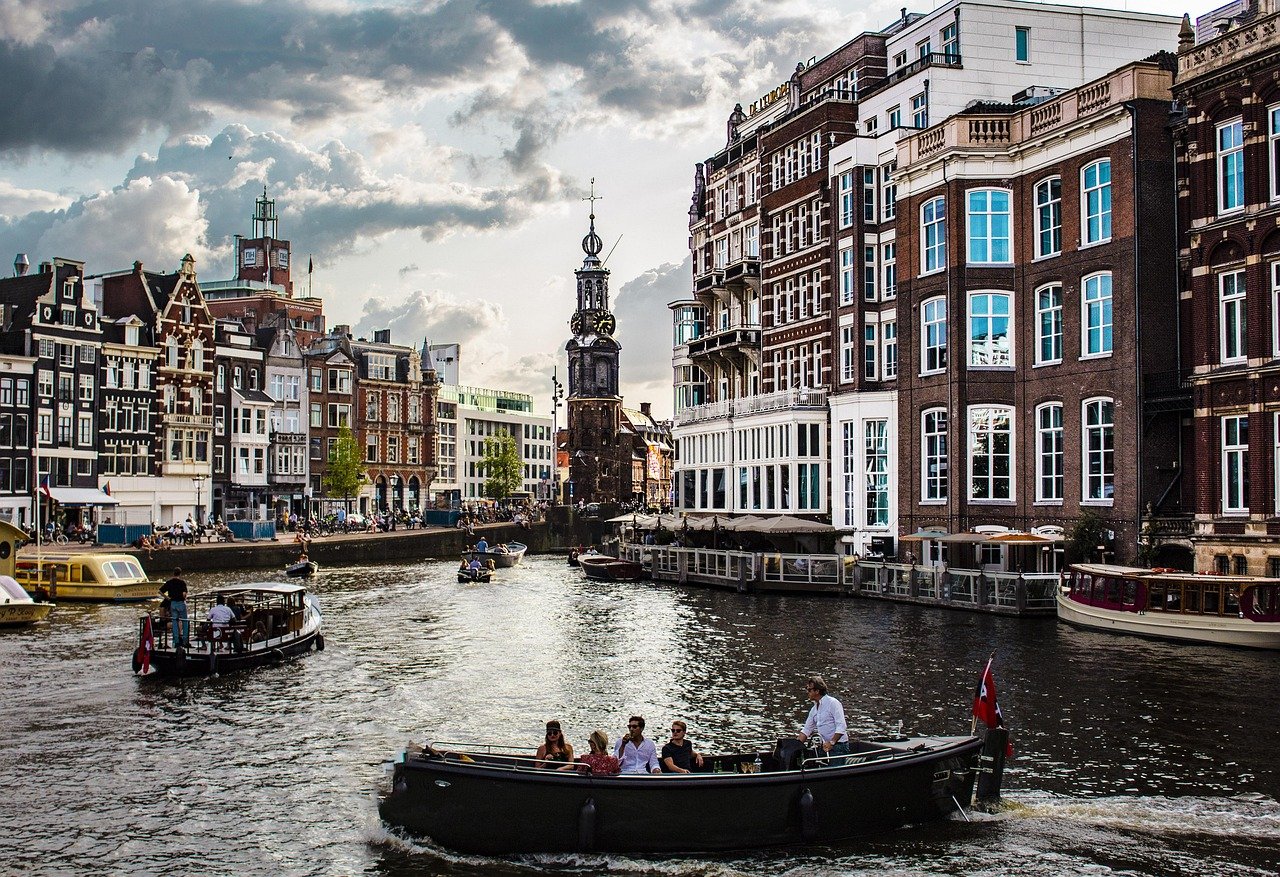 Tourists enjoying boat rides on a canal surrounded by historic buildings in Amsterdam under a cloudy sky