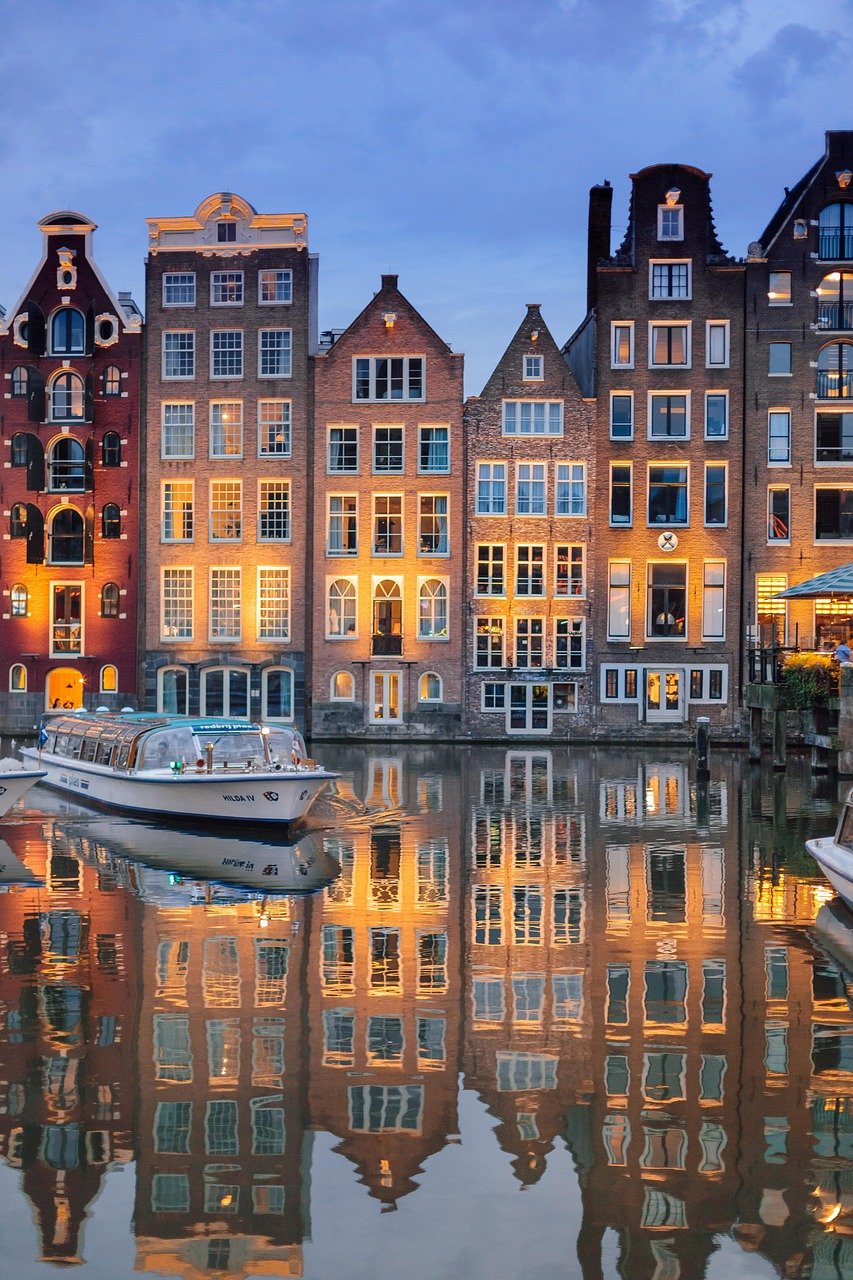 Historic canal houses with illuminated windows reflecting on water in Amsterdam during twilight with a boat in the foreground