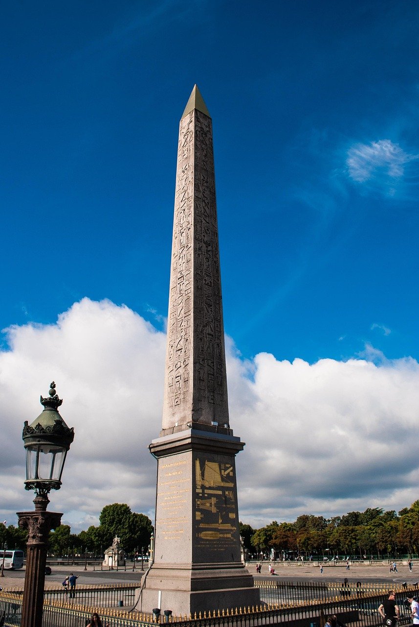 Tall ancient Egyptian obelisk with hieroglyphs in a public square under blue sky and white clouds