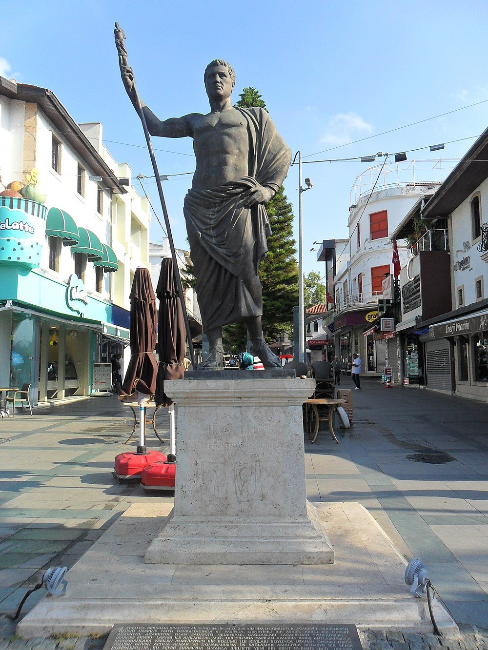 Statue of ancient Greek figure holding a staff in a busy urban pedestrian street with shops and cafes