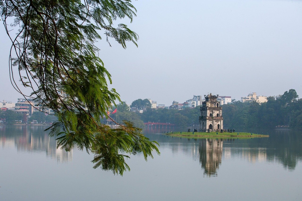 Ancient tower on small island in calm lake with cityscape and green trees in background under clear sky