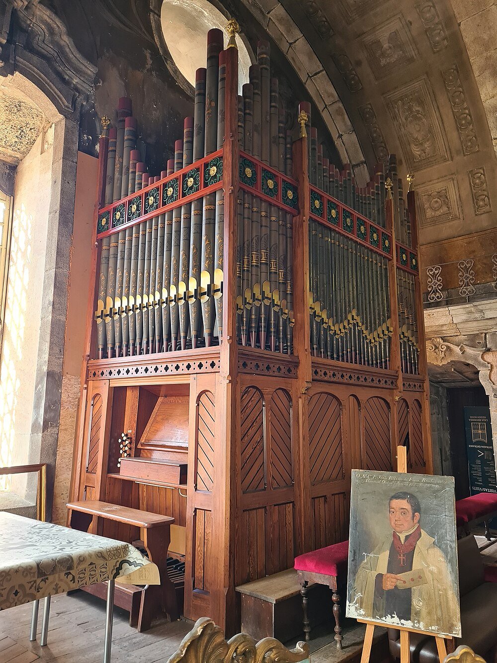 Large antique pipe organ with wooden frame and decorative pipes inside historic stone building with portrait painting nearby