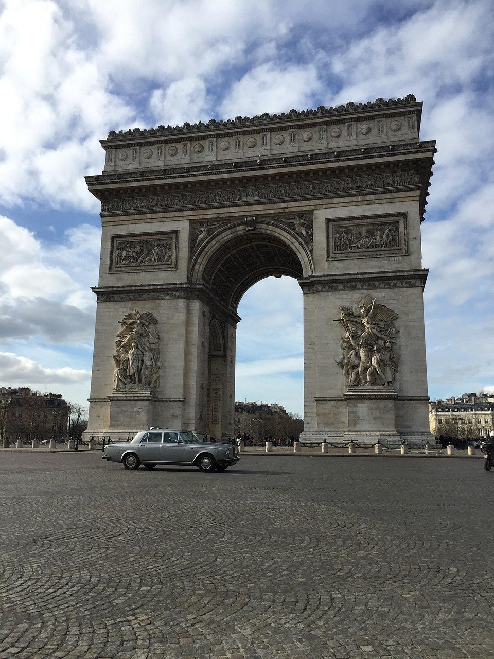 Arc de Triomphe in Paris with a vintage silver car parked in front on a cobblestone plaza under a partly cloudy sky