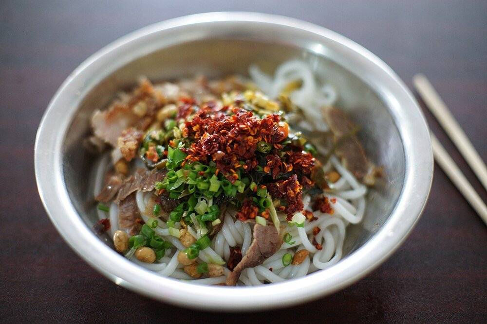 Bowl of Asian rice noodles topped with sliced beef, chopped green onions, peanuts, and red chili flakes with chopsticks nearby