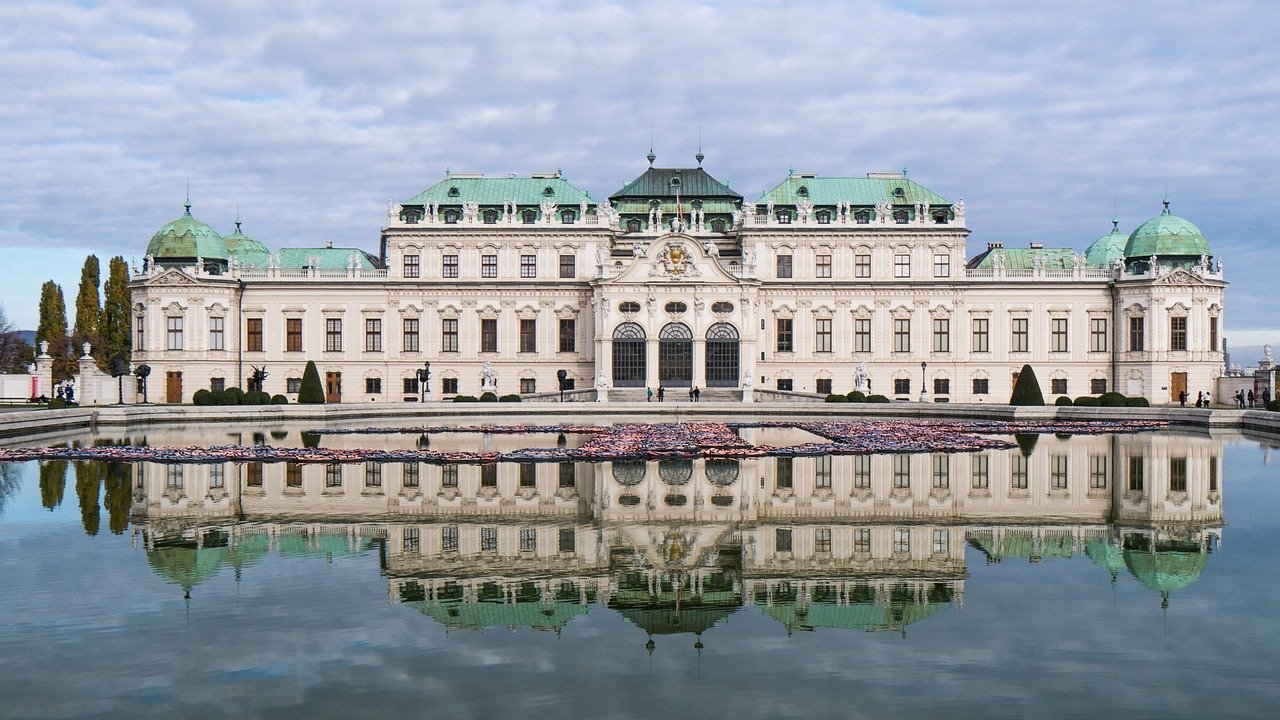 Baroque palace with green domes reflected in a large pond under a cloudy sky, showcasing historic architecture.