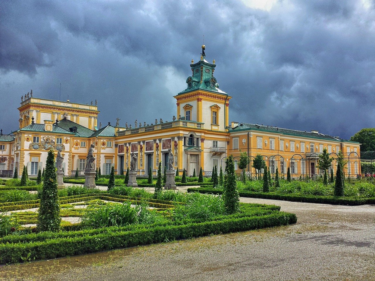 Baroque style palace with ornate statues and manicured gardens under a dramatic cloudy sky