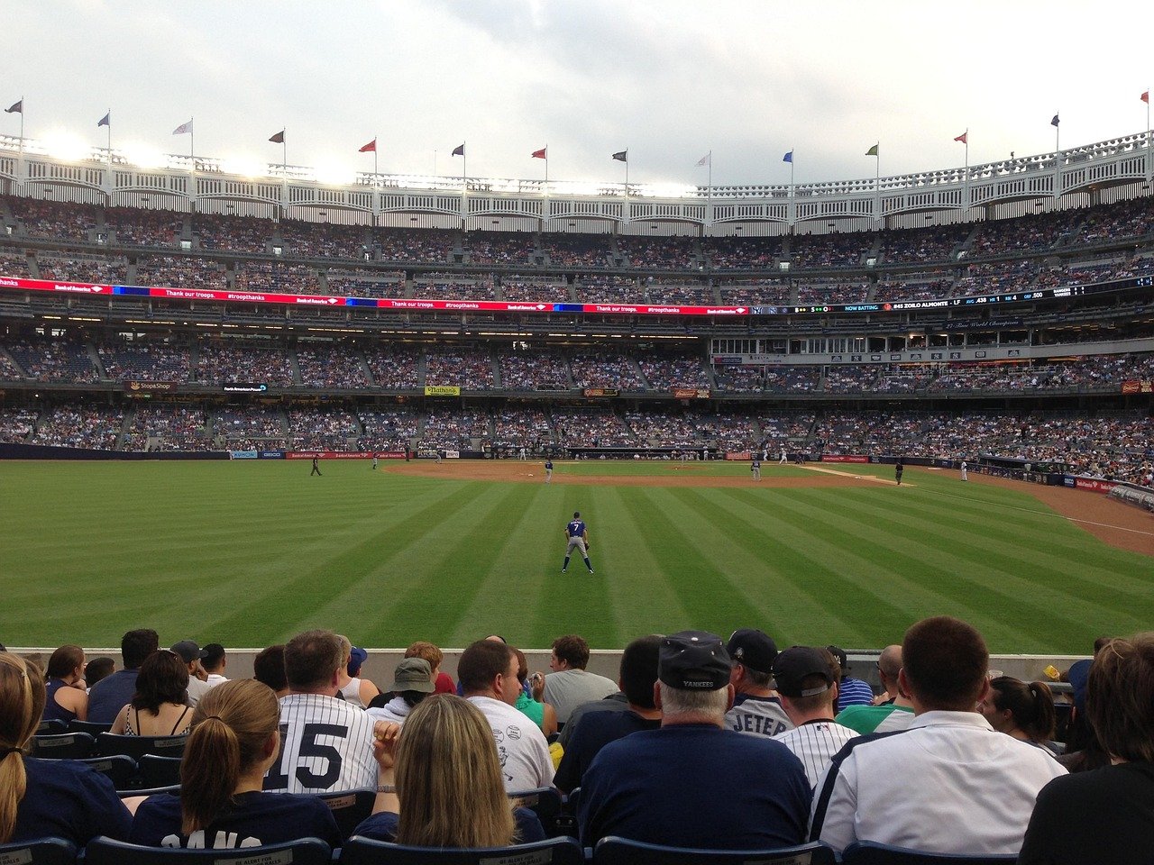 Crowded baseball stadium with fans watching a game on a sunny day, players on the field, and flags flying above stands