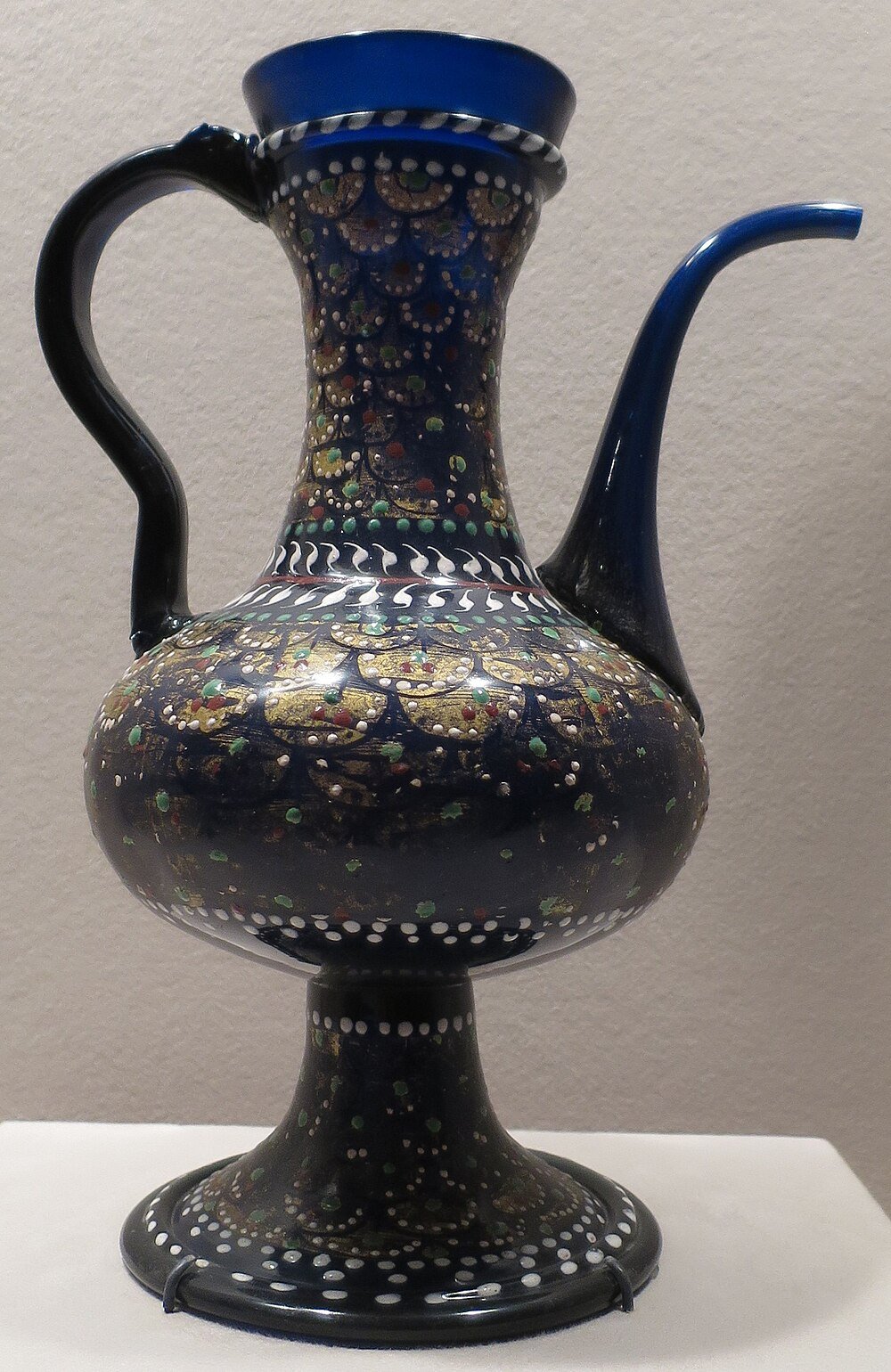 Decorative blue ceramic ewer with intricate gold, green, and red patterns, featuring a curved handle and spout on a pedestal base