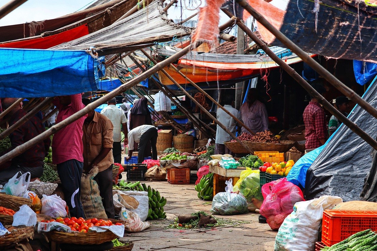 Busy outdoor market with fresh vegetables, fruits, and people shopping under colorful tarps in a vibrant street setting