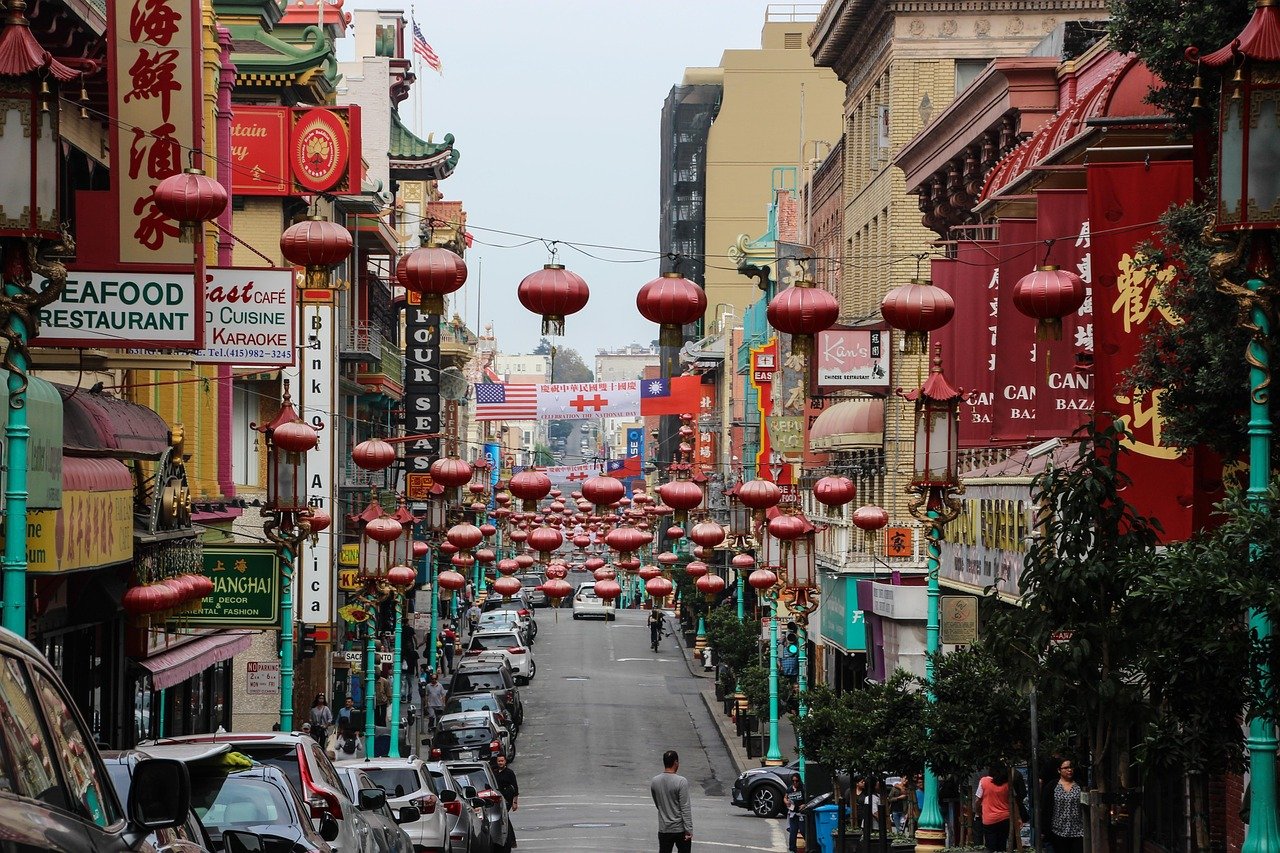 Busy Chinatown street with red lanterns, Asian restaurants, and parked cars in an urban city setting.