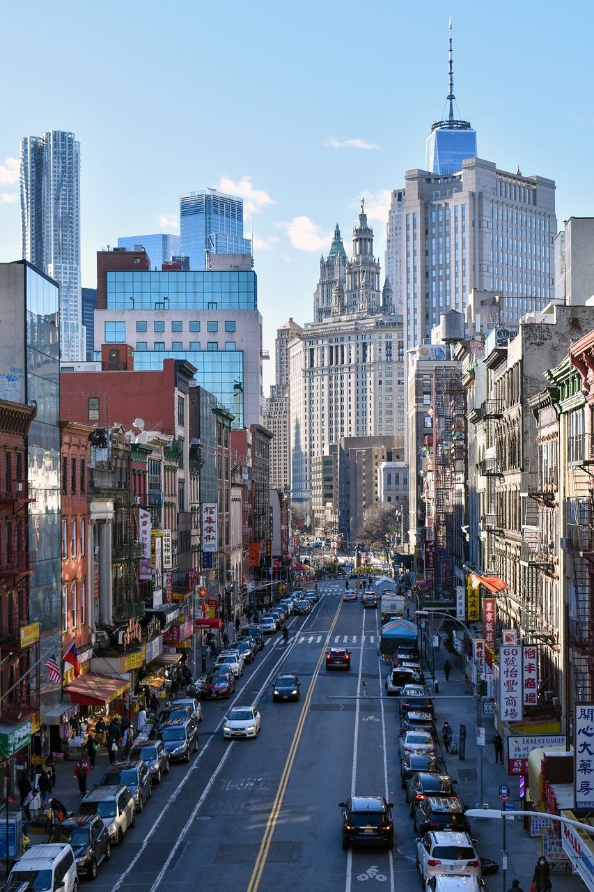 Busy urban street in Chinatown with parked cars, pedestrians, and tall skyscrapers in the background under a clear sky