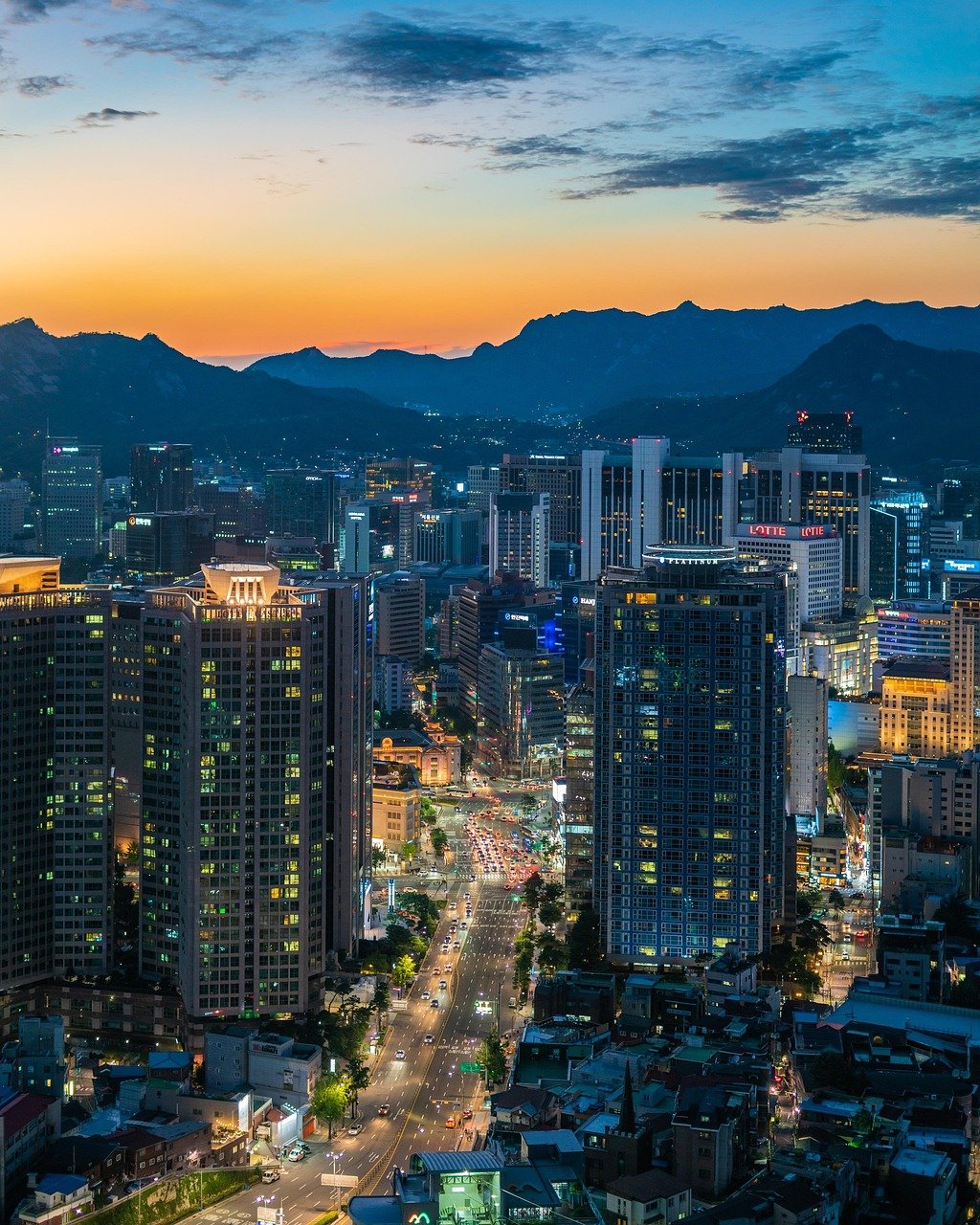 City skyline at dusk with illuminated skyscrapers, busy streets, and mountains in the background under a colorful sky