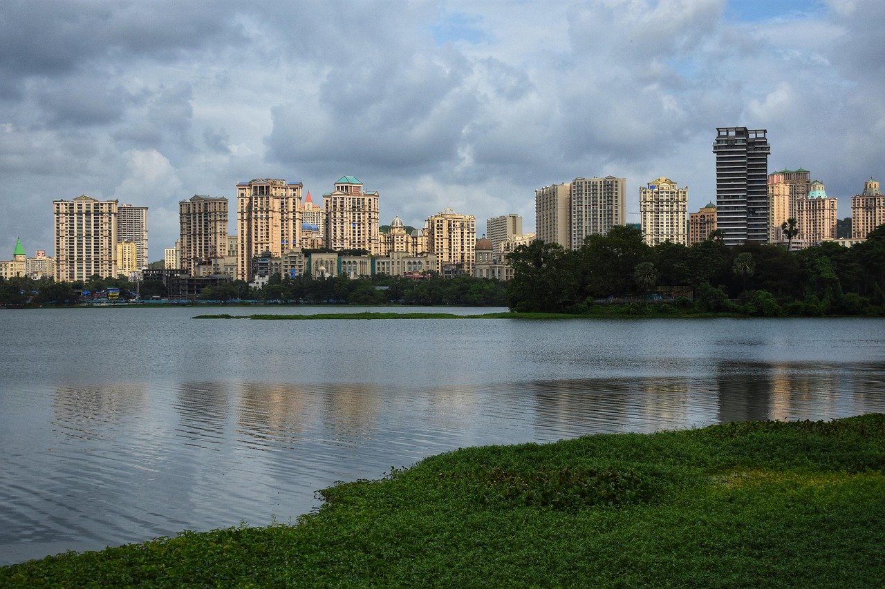 City skyline with high-rise buildings reflected in a calm lake under a cloudy sky with green vegetation in the foreground