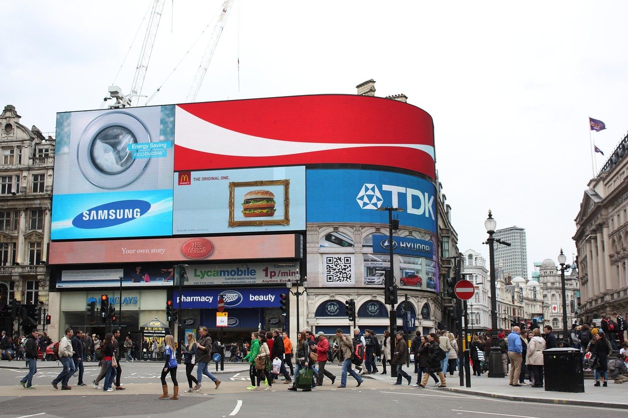 Busy city street with large digital billboards displaying Samsung, McDonald's, TDK, and Hyundai ads, and pedestrians crossing
