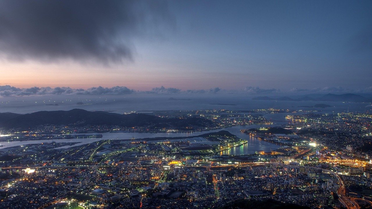 Aerial view of a coastal city at dusk with illuminated buildings, waterways, and distant mountains under a cloudy sky