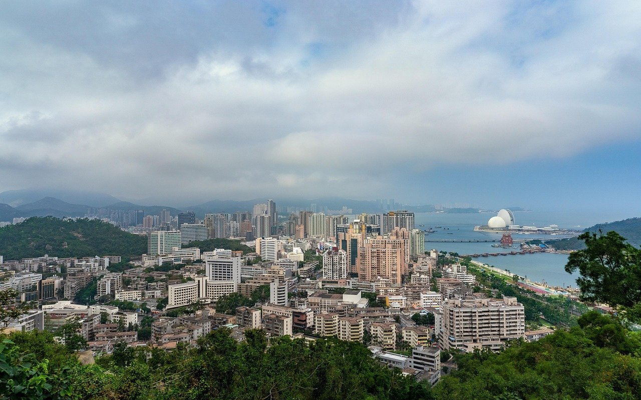 Panoramic view of a coastal city with high-rise buildings, green hills, and a harbor with unique dome-shaped structures under a cloudy sky
