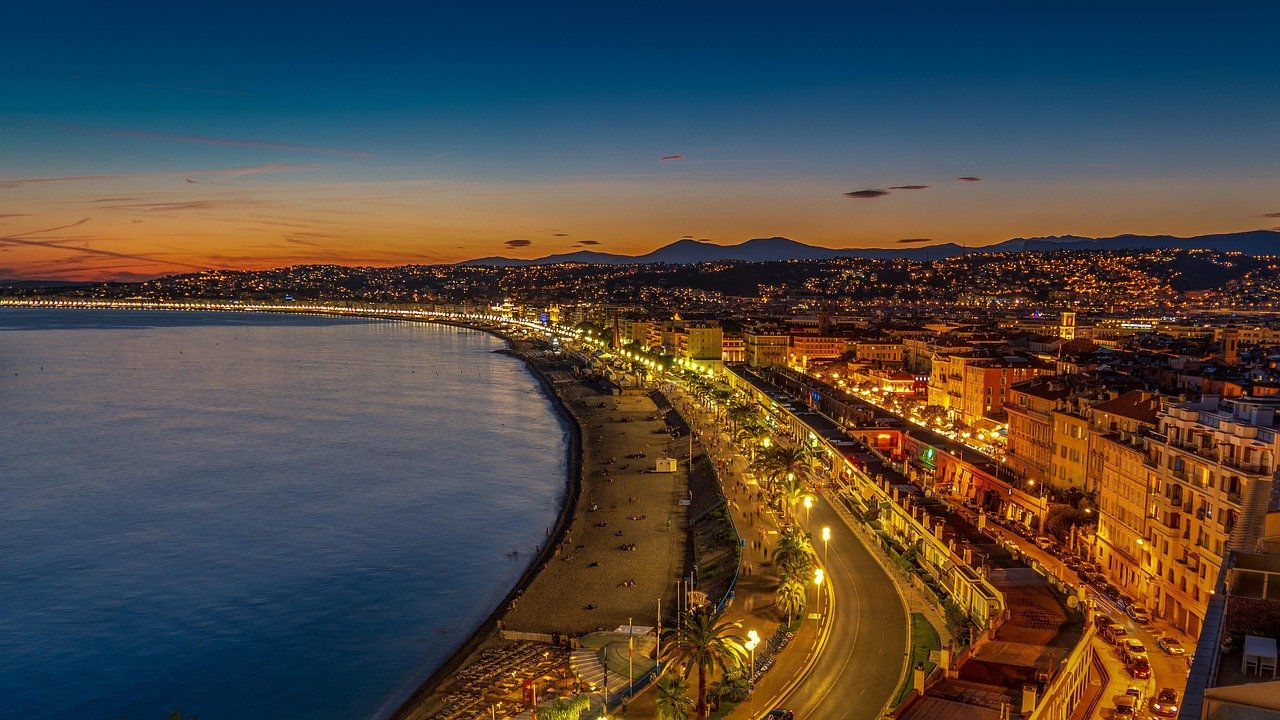 Nighttime view of a coastal city with illuminated streets, beach, and mountains in the background at sunset