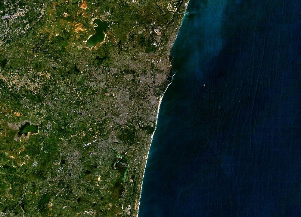 Satellite view of a coastal city with urban areas, green landscapes, and ocean coastline, showing detailed land and water features.