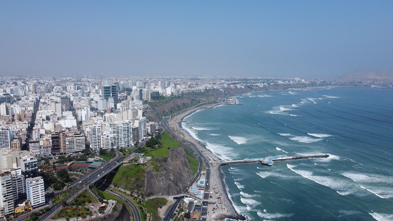Aerial view of a coastal city with high-rise buildings, cliffs, a beach, and ocean waves under a clear sky