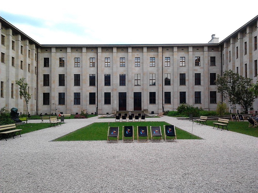 Outdoor courtyard with gravel paths, green lawn, benches, and deck chairs in front of a large historic building with multiple windows