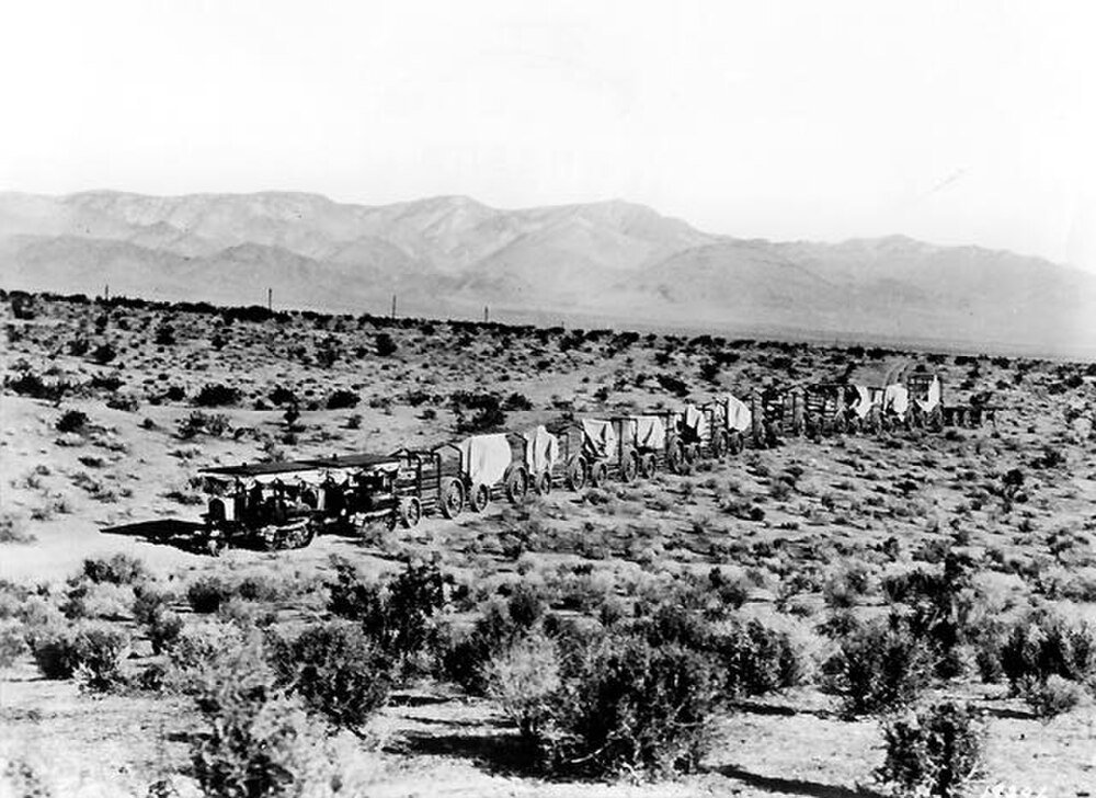 Historic black and white photo of a long line of covered wagons crossing a desert landscape with mountains in the background