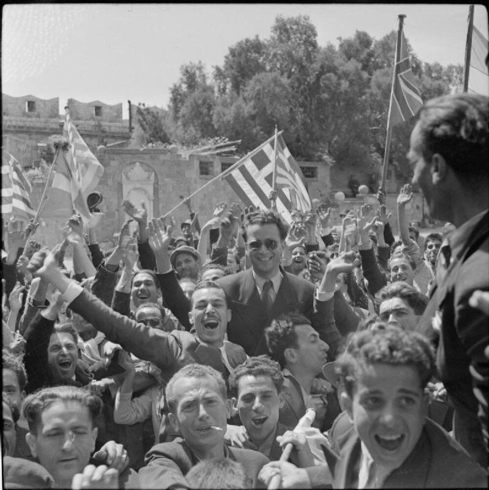 Jubilant crowd waving flags outdoors Crowd of jubilant men waving flags and celebrating outdoors in a historic setting with stone buildings and trees