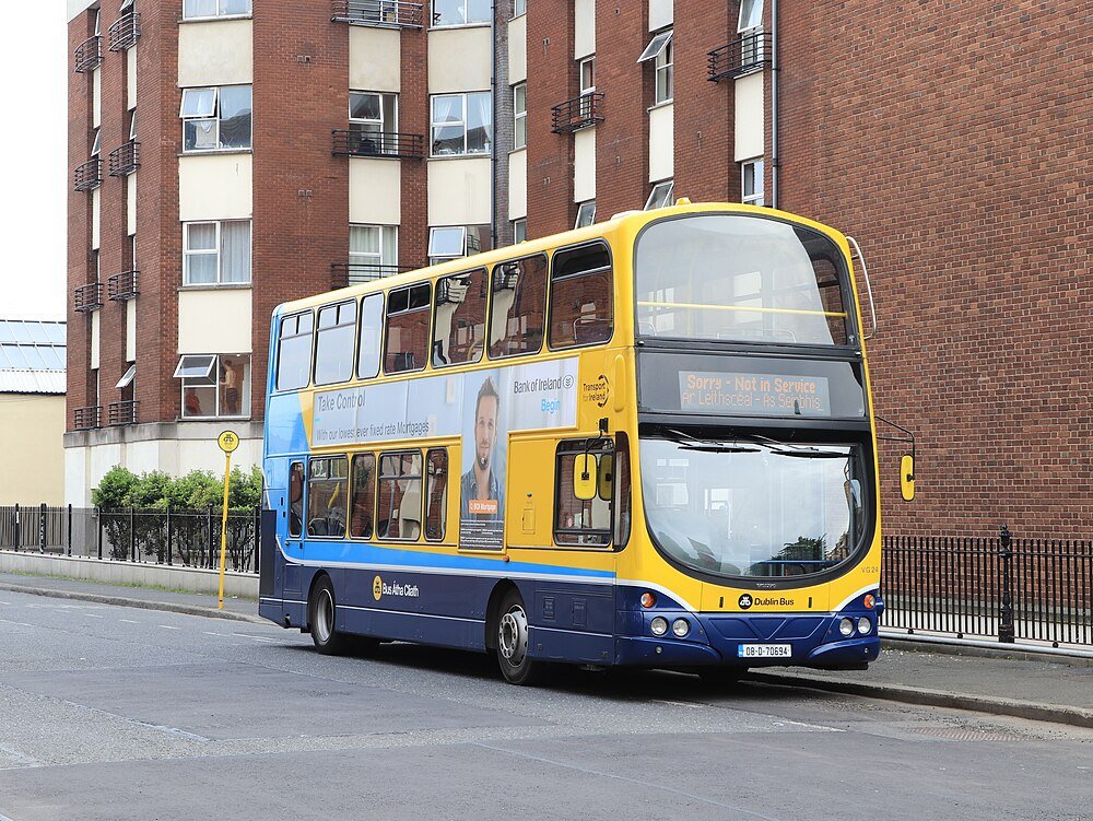 Double-decker Dublin bus parked on city street with brick buildings and advertisement on side panel