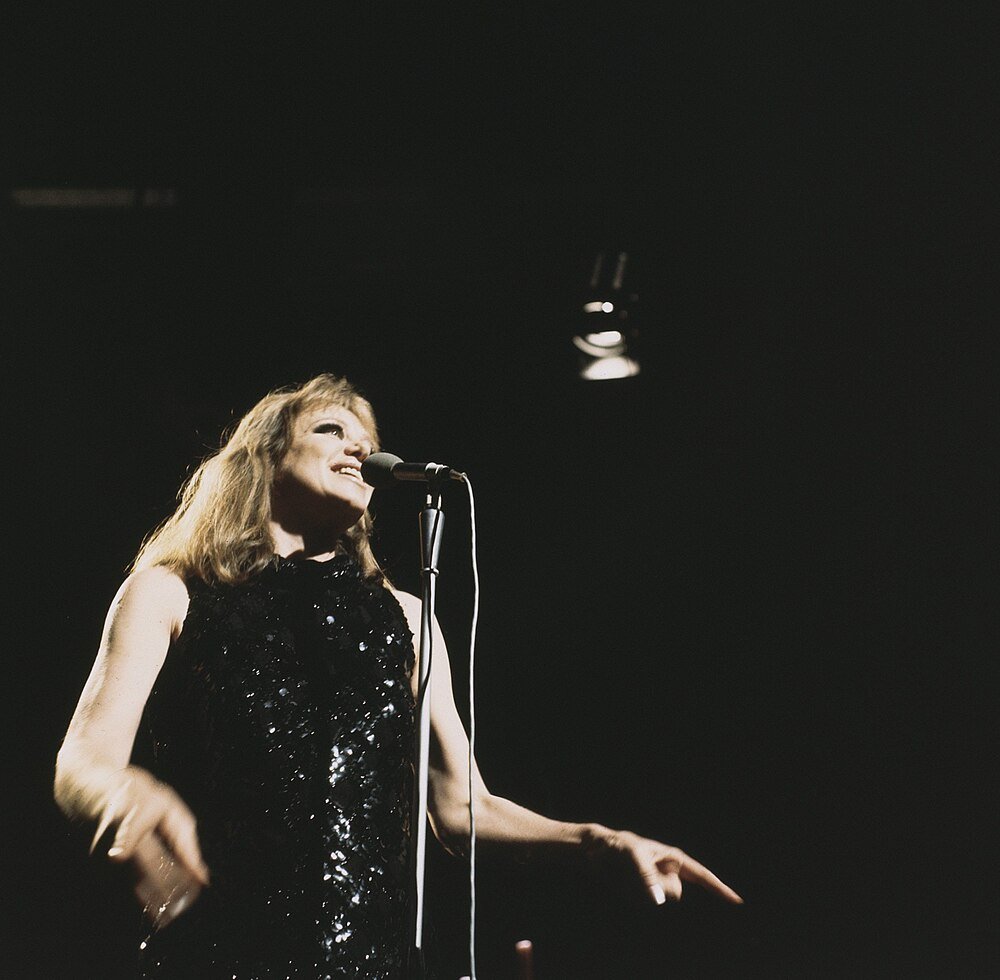Female singer performing on stage with microphone in a black sequined dress under spotlight