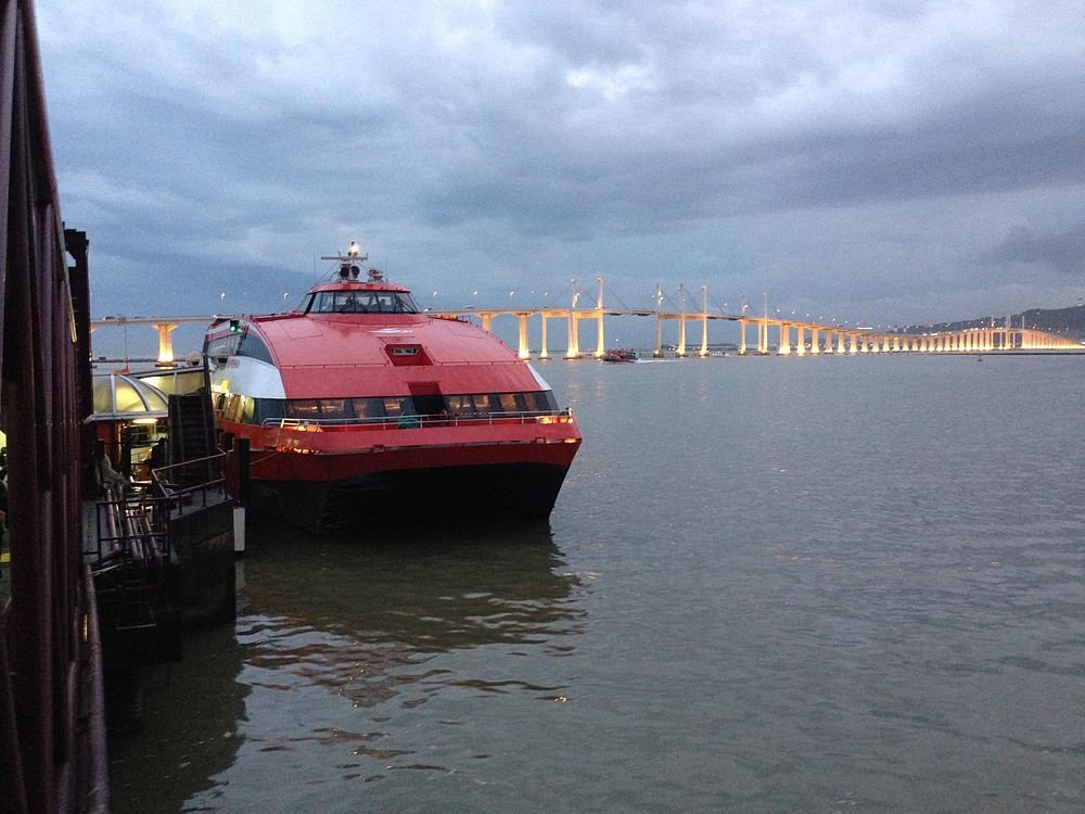 Red and black ferry boat docked near a pier with a long illuminated bridge in the background during dusk