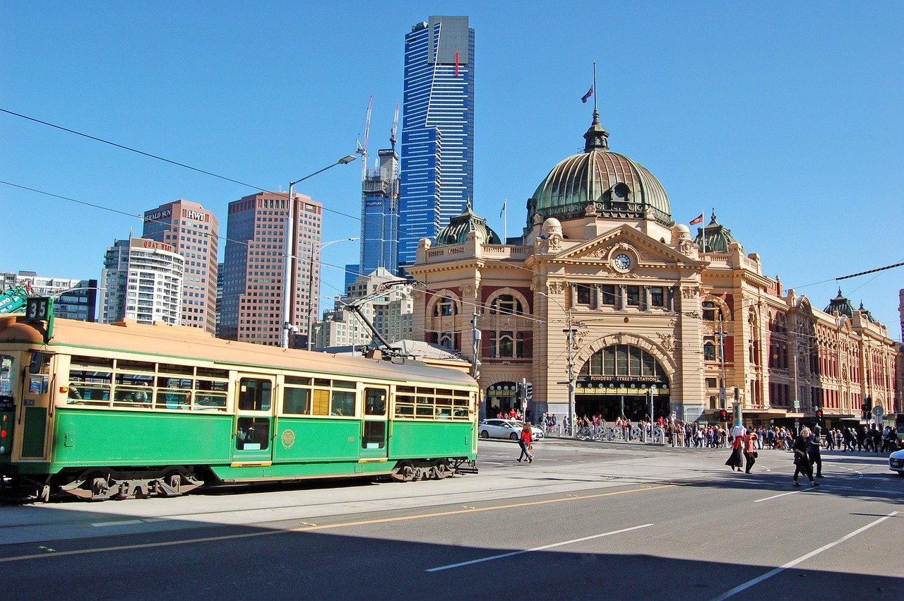 Historic Flinders Street Station with green tram and modern skyscrapers in Melbourne city under clear blue sky