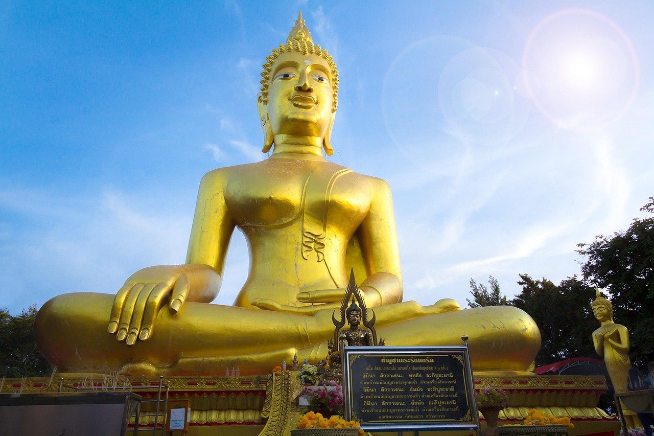 Large golden Buddha statue seated outdoors under blue sky with smaller statues and Thai script plaque in front