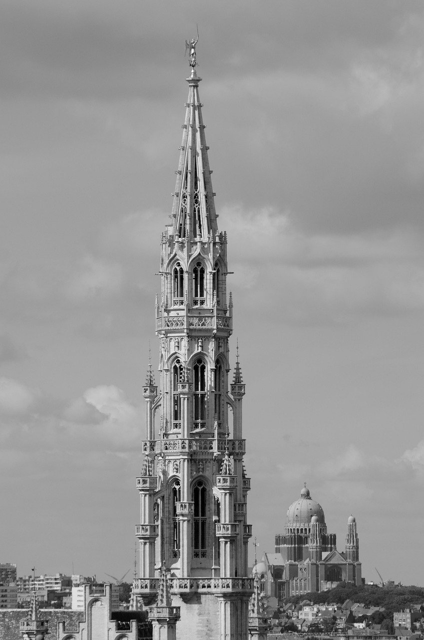 Gothic-style tall church tower with intricate details and a statue on top under cloudy sky in an urban setting