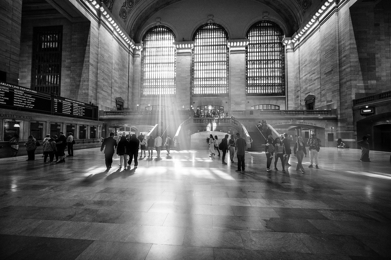 Black and white image of Grand Central Terminal with sunlight streaming through large windows and people walking inside