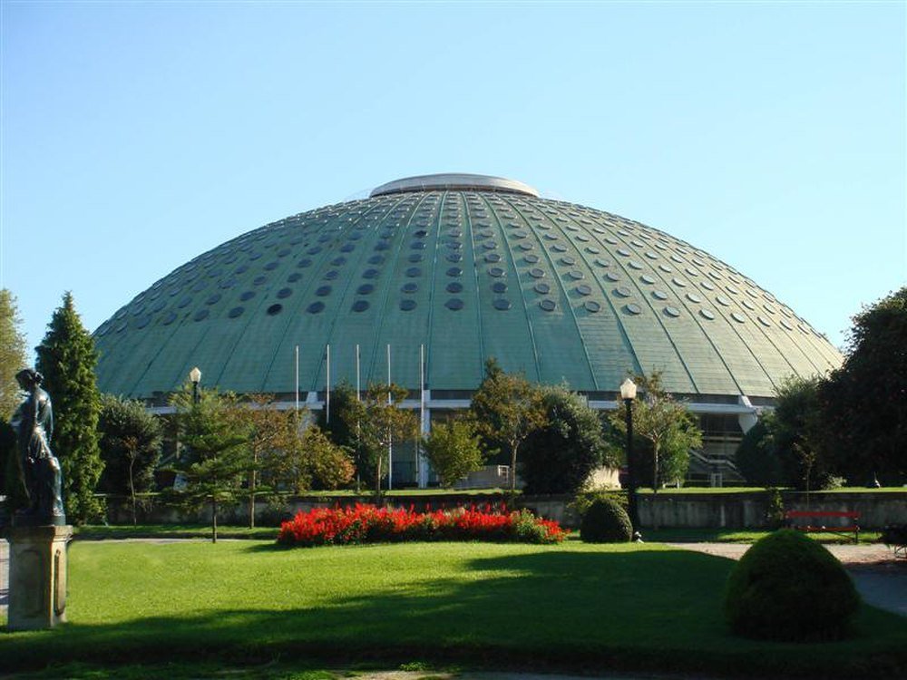 Green dome building with circular windows and garden Large green dome-shaped building with circular windows surrounded by trees and a garden with red flowers