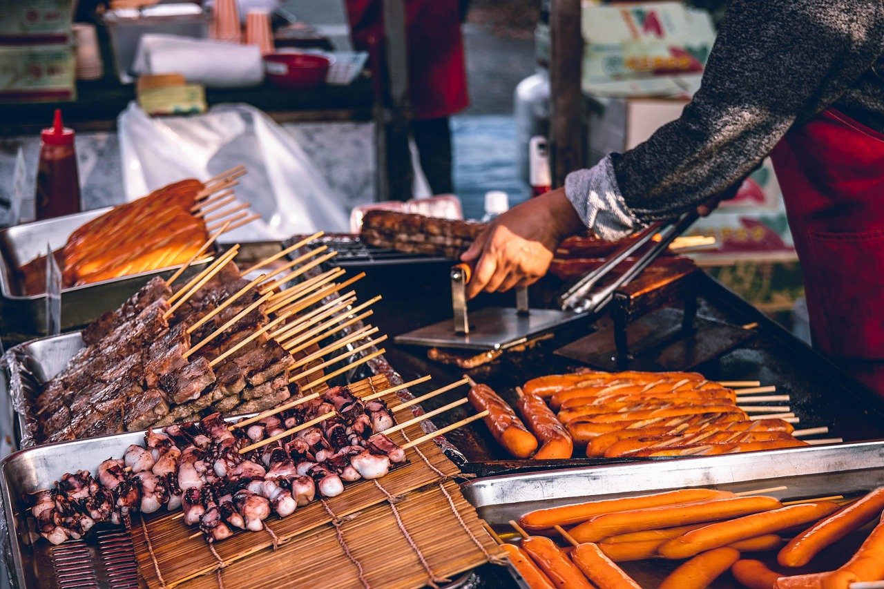 Street food vendor grilling assorted skewered meats and sausages on an outdoor grill at a busy market stall.