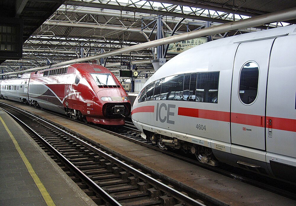 Two modern high-speed trains at a busy train station platform with overhead metal roof and tracks.