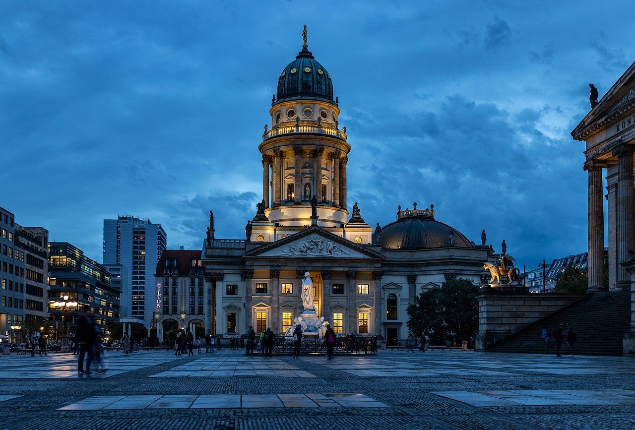 Illuminated historic cathedral with dome and statues in urban square at dusk with people walking around