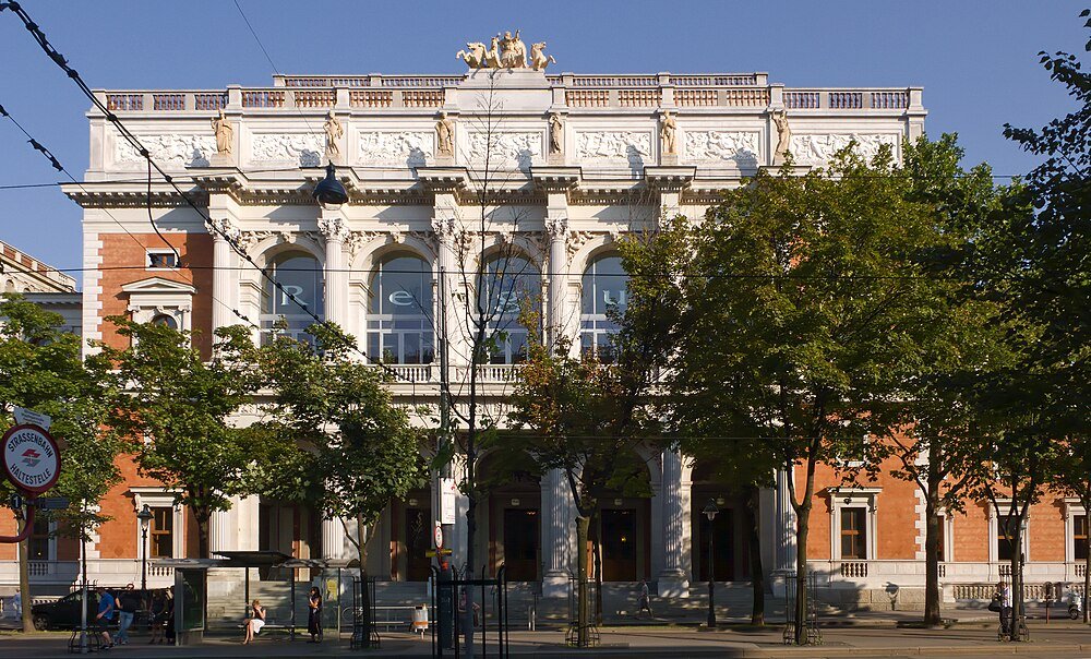 Historic classical building with sculptures and trees Historic classical building with ornate sculptures and large arched windows behind green trees on a sunny day