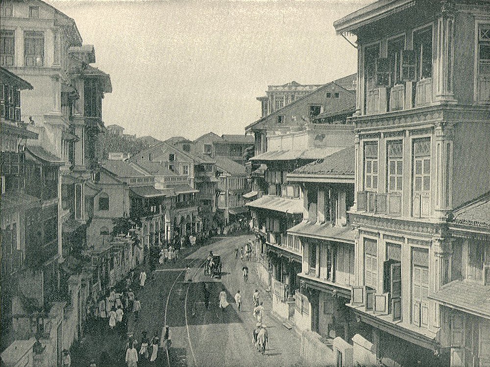 Historic black and white photo of a busy street with colonial-style buildings and people walking and riding horses