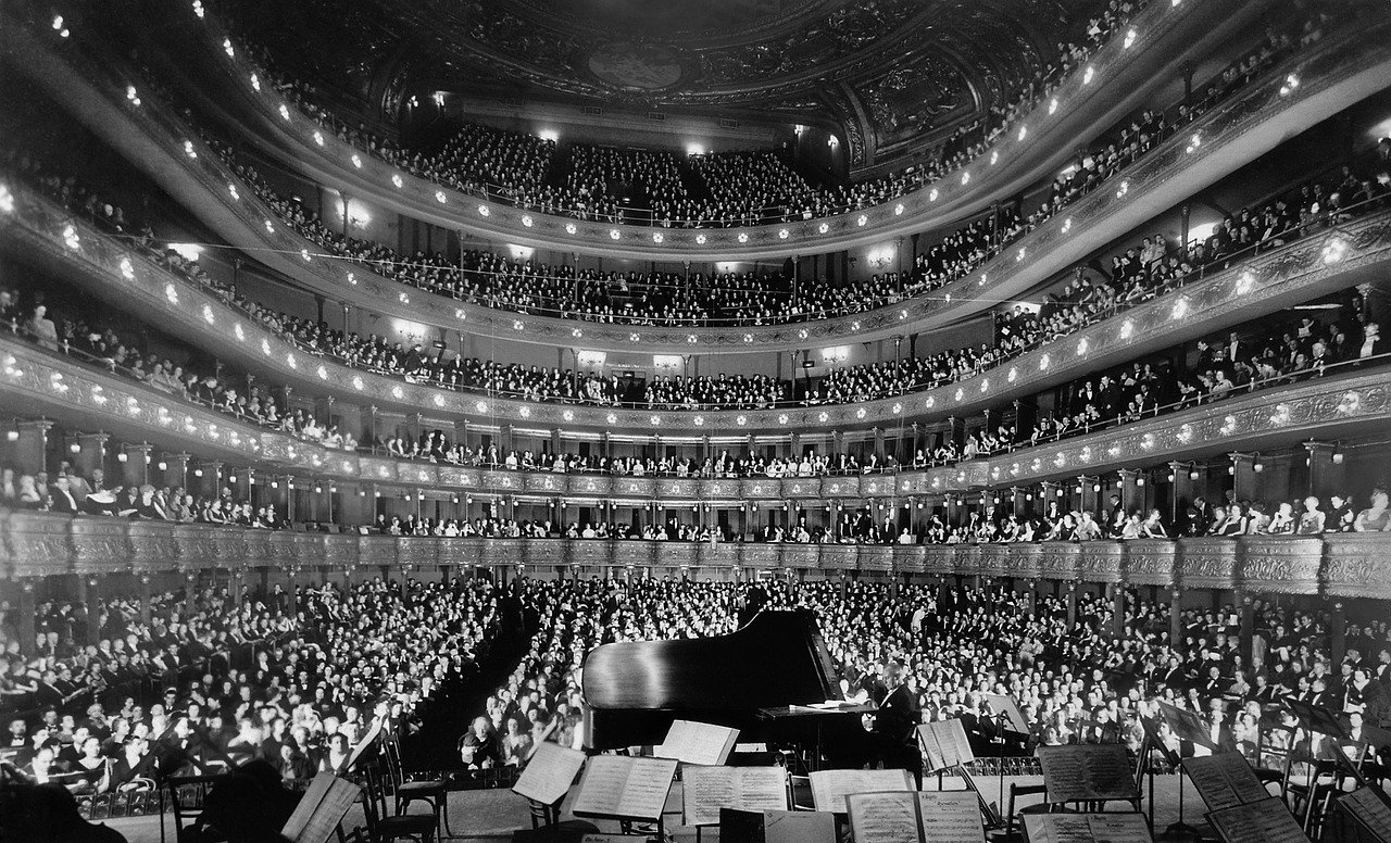 Large audience in a grand historic concert hall watching a pianist perform on stage with orchestra music stands