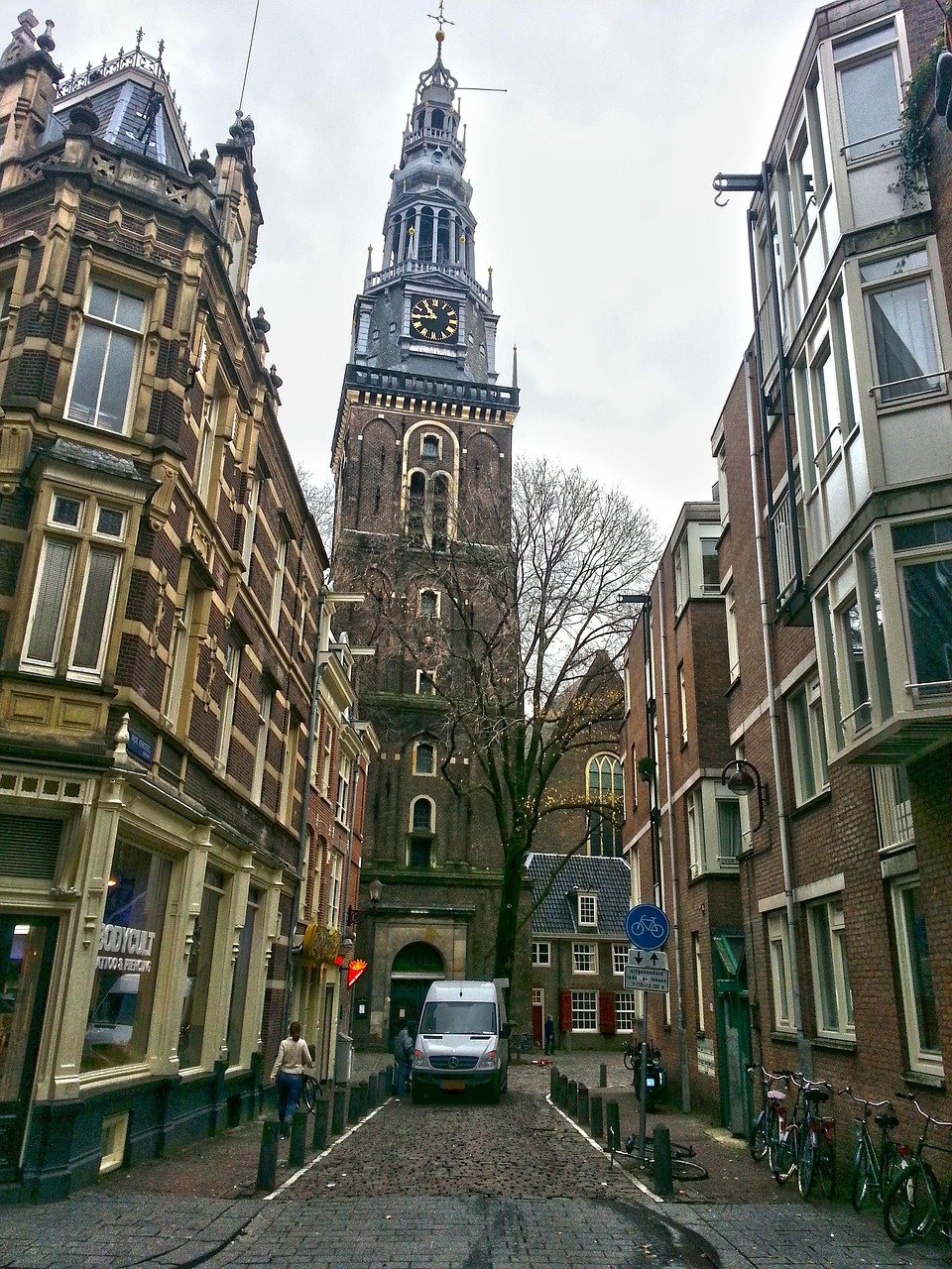 Historic European street with cobblestone road, old brick buildings, bicycles, and a tall clock tower under cloudy sky