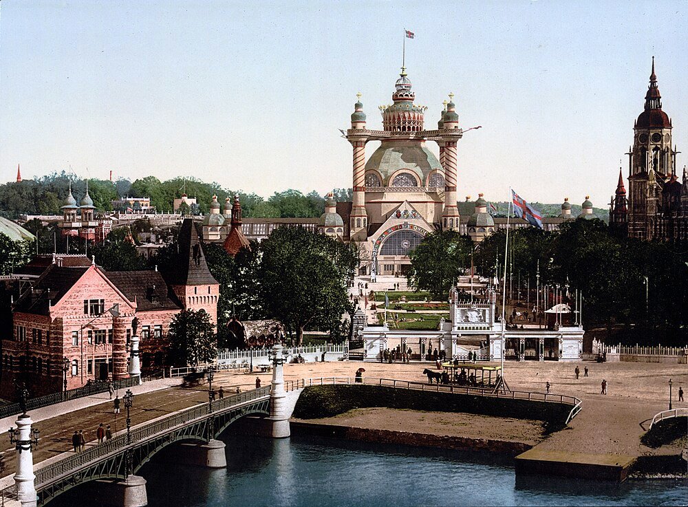 Historic view of a grand exhibition hall with ornate architecture, a bridge over water, and surrounding buildings in a city park.