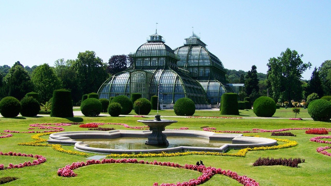 Historic glass greenhouse with ornate fountain and manicured garden featuring colorful flower beds and trimmed bushes