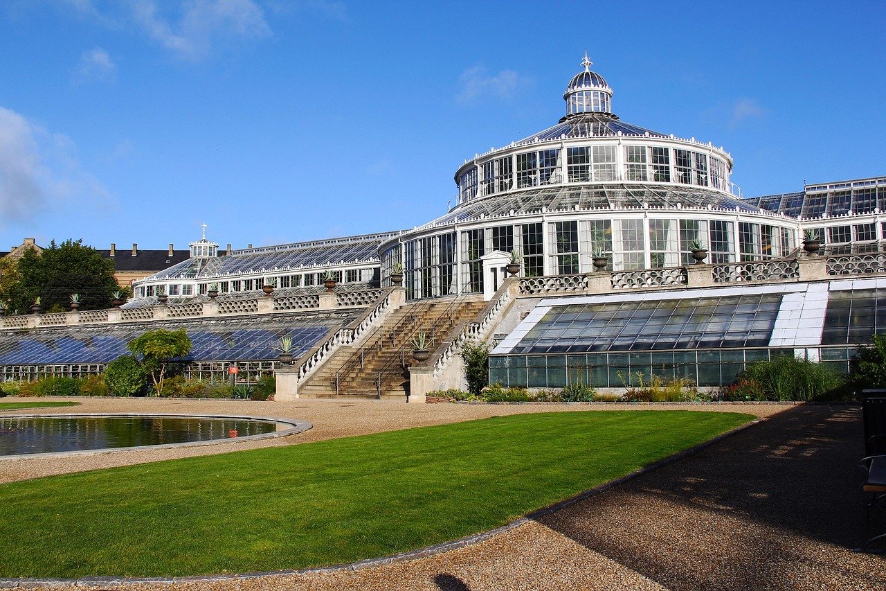 Large historic glass greenhouse with tiered glass roofs and stone stairs in a botanical garden under blue sky