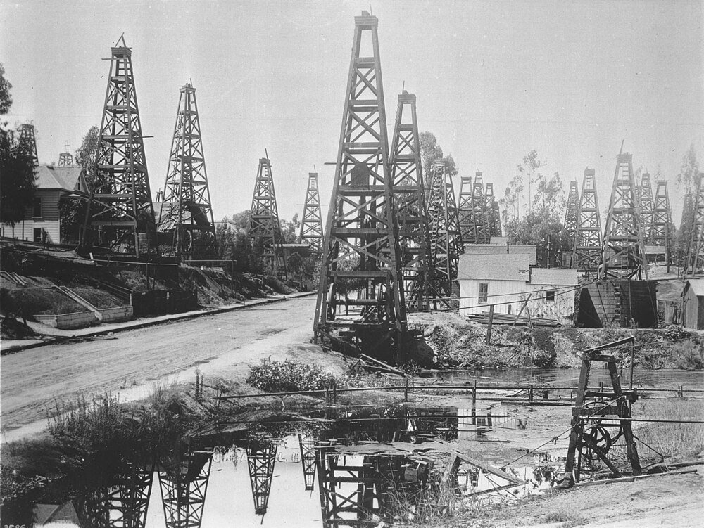 Historic black and white photo of early 20th century oil drilling rigs and wooden derricks near a water body